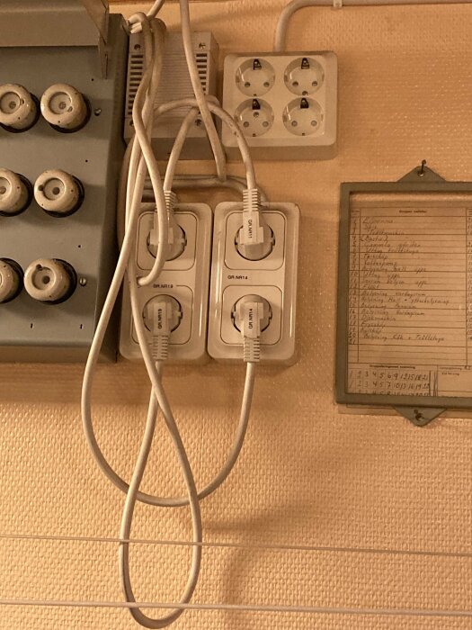 Väggmonterade elektriska uttag med inkopplade kablar, åldrad dokumentram och retro strömbrytare på tapetserad vägg.