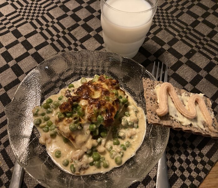 Middagstallrik med gröna ärter, gräddsås, stekt fiskfilé, knäckebröd med pålägg, mjölk, rutig bordstablett.