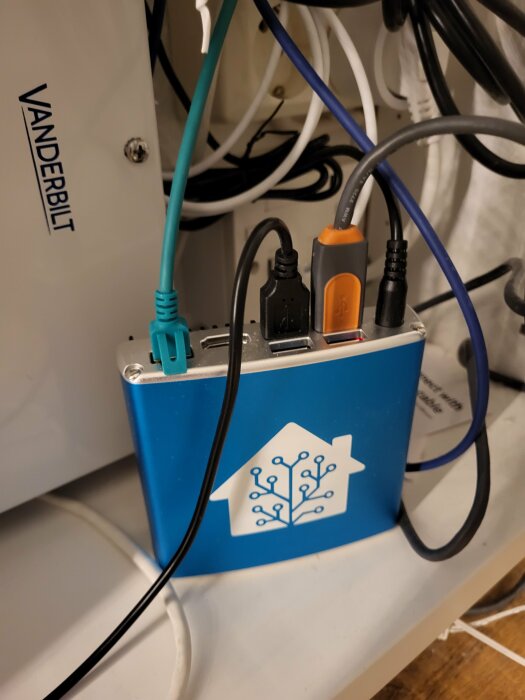 Elektronisk enhet med kablar och logotyp på hylla, blandat kabeltrassel i bakgrunden.