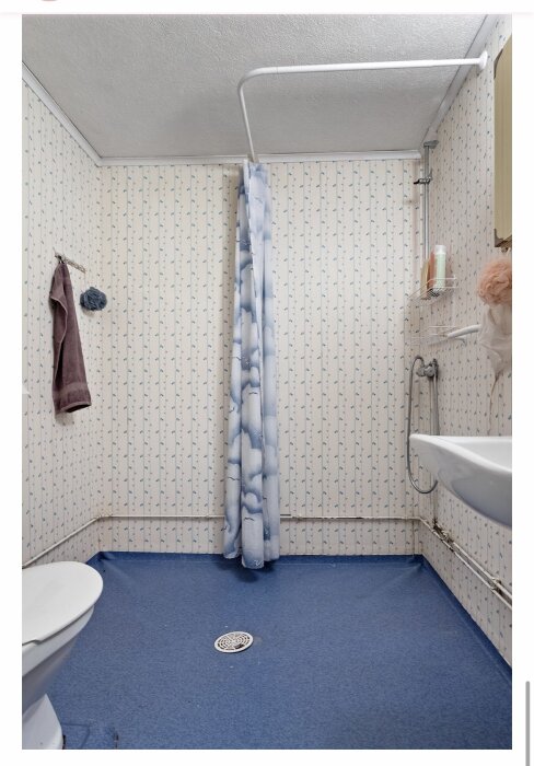 Ett gammaldags badrum med badkar, dusch, toalett, blåmönstrad matta och blommigt kakel.