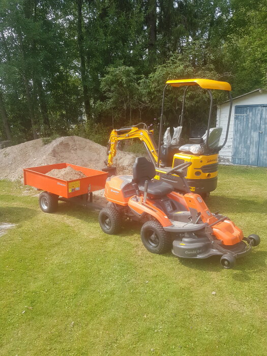 Orange trädgårdstraktor kopplad till släpvagn på gräs framför hög med grus och skogsbakgrund.