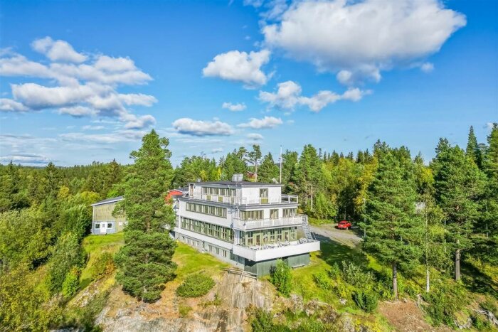 Stort hus på klippa, omgivet av skog, under blå himmel med moln.