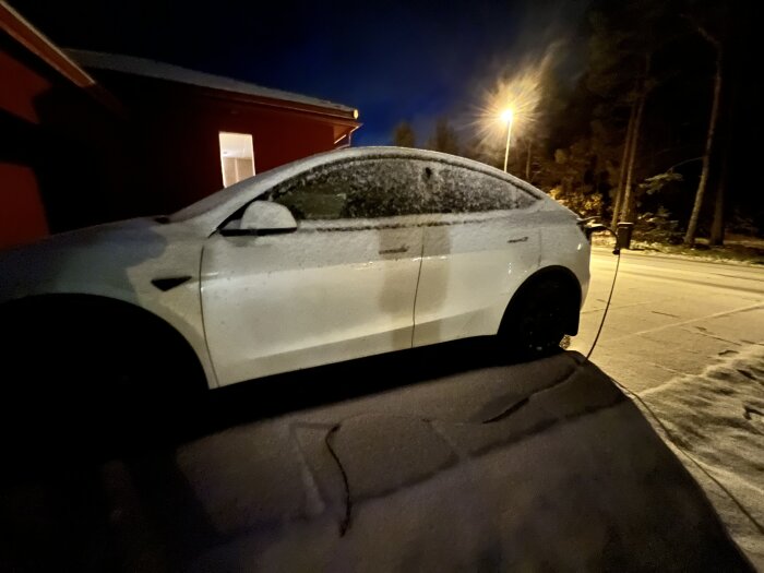 Vit elbil parkerad nattetid, täckt med snö, laddning pågår, vid hus.