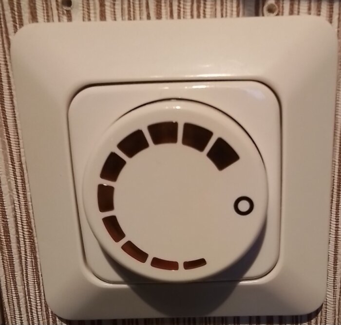 Värmeelementets termostat med vridkontroll, bruna markeringar, monterad på vägg, vit bakgrund.