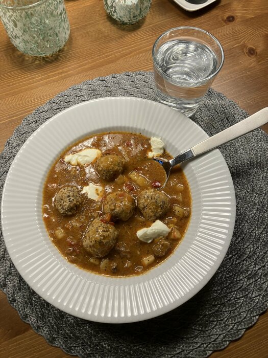 Gryta med köttbullar, sås, vit klump (eventuellt färskost), glas vatten, rutig bordstablett, träbord.