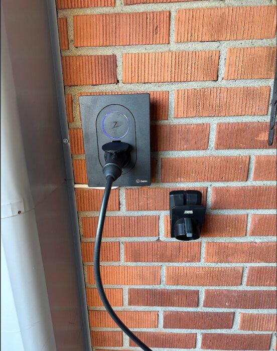 Laddstation för elbil med inkopplad kabel, mot tegelvägg, utomhus, dagtid.