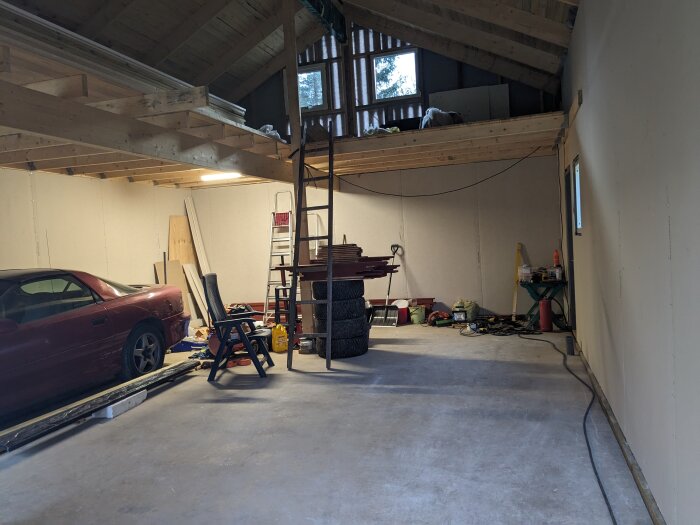 Garage med bil, röra, arbetsbänk, stege, öppet tak. Verktyg och material spridda omkring. Under uppbyggnad eller renovering.