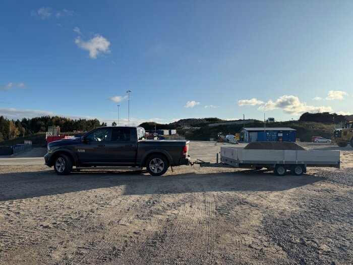 Pickup med släp på grusplan, molnig himmel, byggplats i bakgrunden.