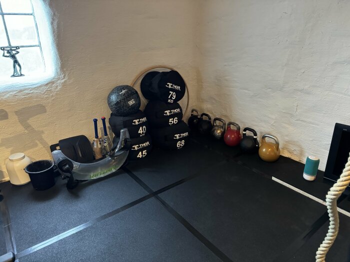 Hemmagym med kettlebells, medicinboll, sandsäckar, roddmaskin och träningstillbehör i ett ljust rum.