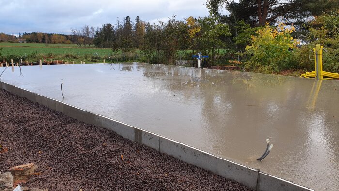 En färsk betongplatta, byggarbetsplats, våt yta, skogsmark, grå moln, hösttid, konstruktionsmaterial runtomkring.