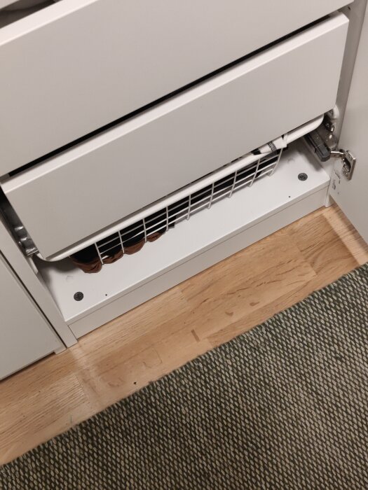 En vit radiator över trägolv och matta med synliga rör och ventilkopplingar.