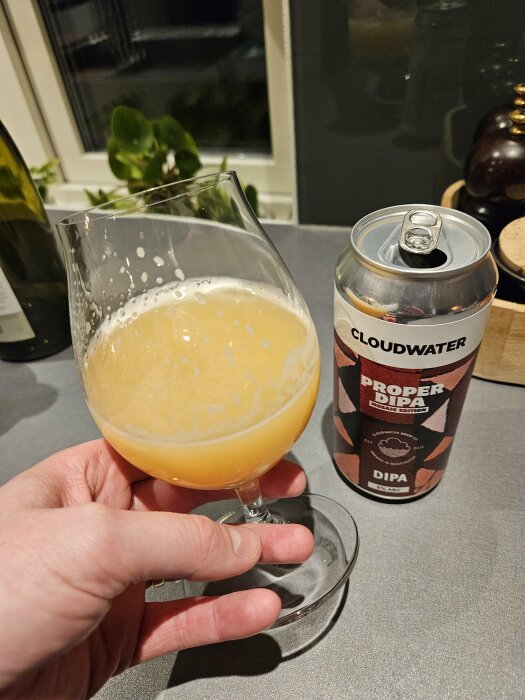 Ölglas med skummande öl, hand håller, ölburk från Cloudwater, texten "Proper DIPA", köksmiljö.