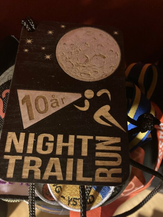 Trämedalj med texten "10 år NIGHT TRAIL RUN", måne och stjärnor, upphängd på färgglada band.