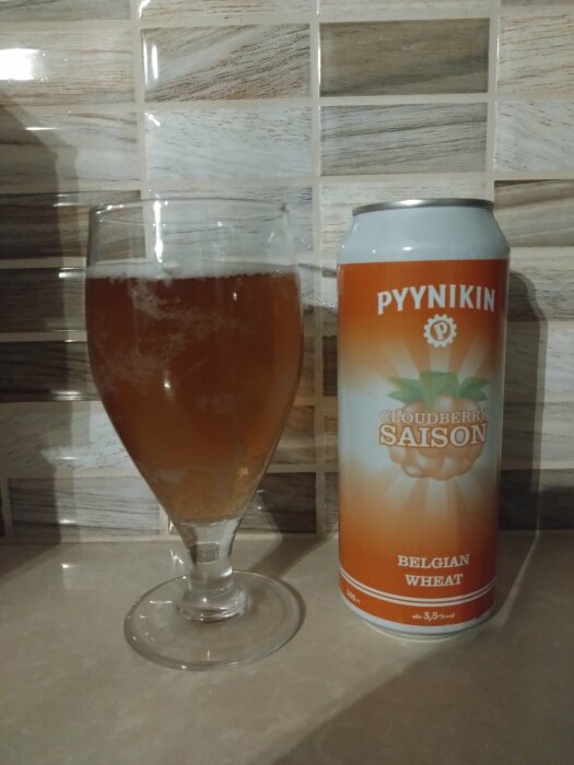 Ett ölglas fyllt med skummande dryck och en ölburk med texten "PYYNIKIN CLOUDBERRY SAISON BELGIAN WHEAT" på kakelbakgrund.