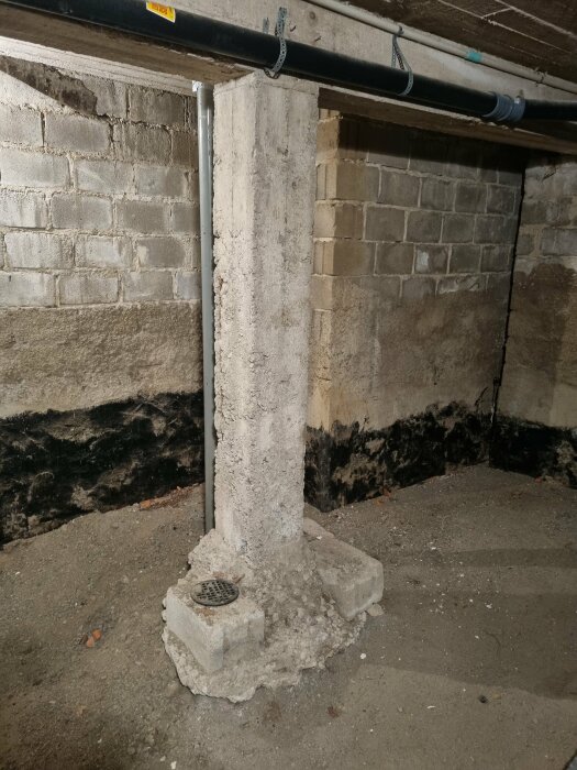 En betongpelare med förstärkning i källare, skadad isolering, rörledningar ovanför, fuktskador på väggarna.