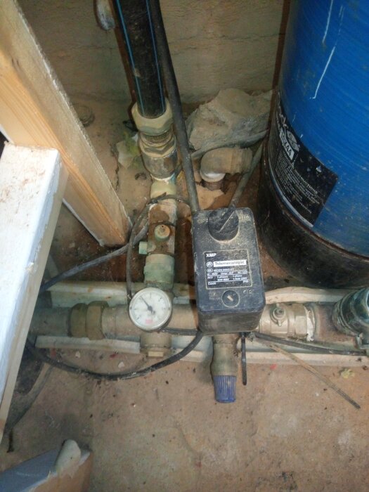 Vattenpump och tryckmätare vid blå behållare i oordnad, dammig miljö.