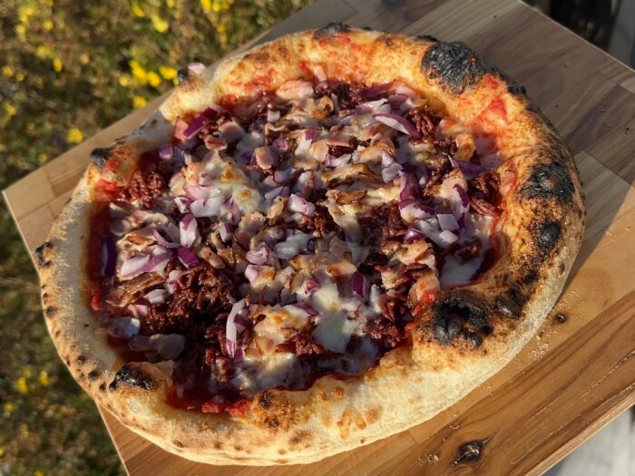Nygräddad pizza med lök och ost på träbräda utomhus i solljus.