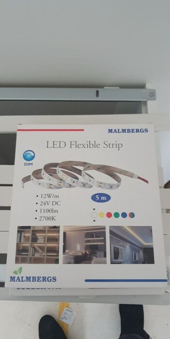 En förpackning med LED Flexible Strip från Malmbergs. Specifikationer och exempelbilder visas. Inomhusmiljö, personens skor syns nedtill.