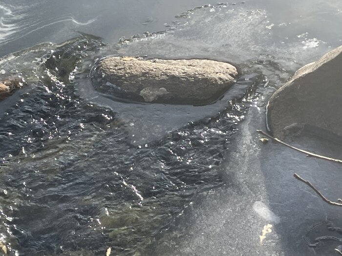 Stenar i rinnande vatten, delvis isbelagt, glittrande solljus, naturlig utomhusmiljö, lugnande landskap.