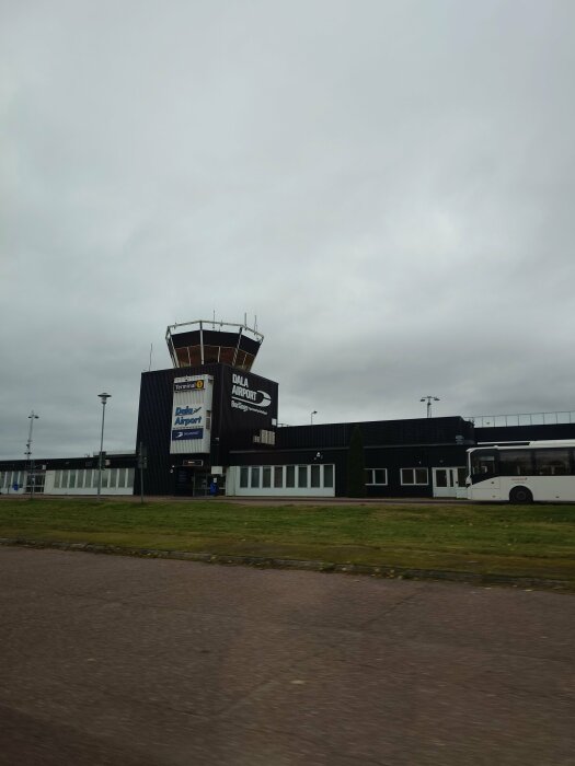 Flygplatsterminal med kontrolltorn, buss, molnig himmel, gräsplan, skyltar med text "Dala Airport" och "Terminal".