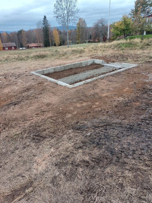 Betongfundament på bar mark, höstträd, röda stugor i bakgrunden, öppet landskap, mulen himmel.