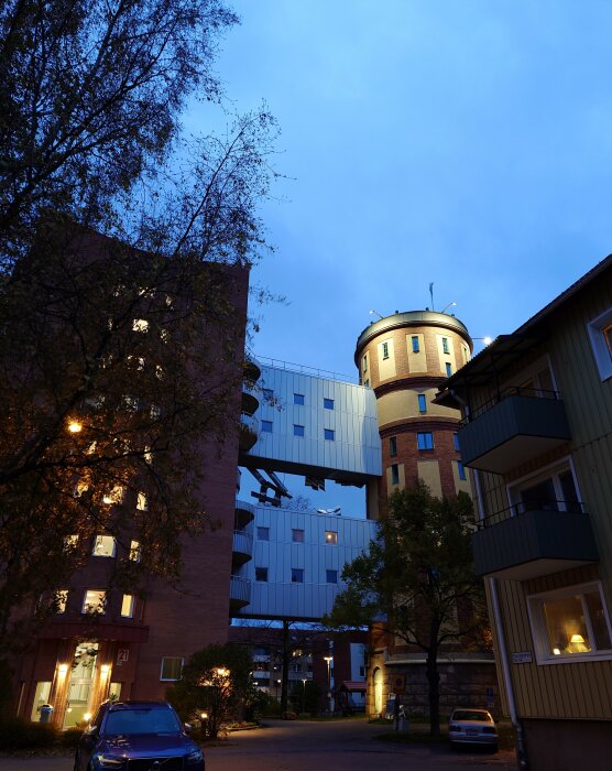 Kvällsstadsscen med blandade byggnadsstilar, bostadshus, vattentorn, blå himmel och tända gatulampor.
