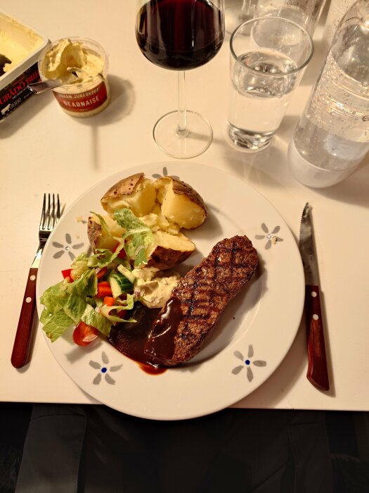 Middag med grillad stek, bakad potatis, sallad, bearnaisesås, rödvin och vatten. Matuppläggning på blommönstrad tallrik.