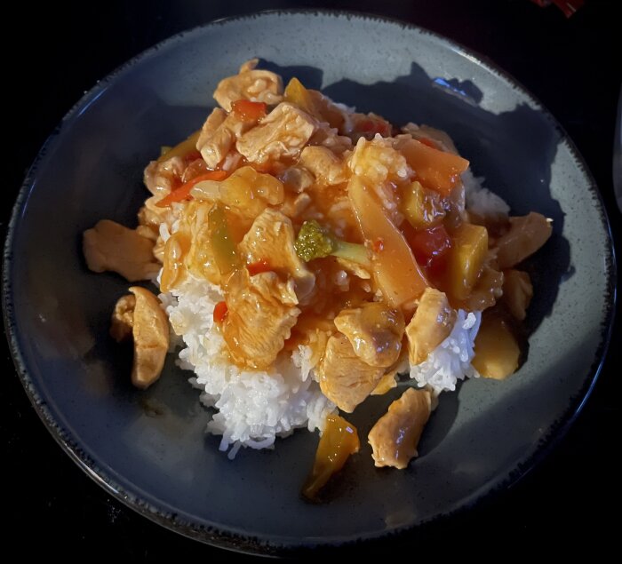 Kyckling i sötsur sås serverad på ris i en grå tallrik. Vibranta färger, asiatisk maträtt.