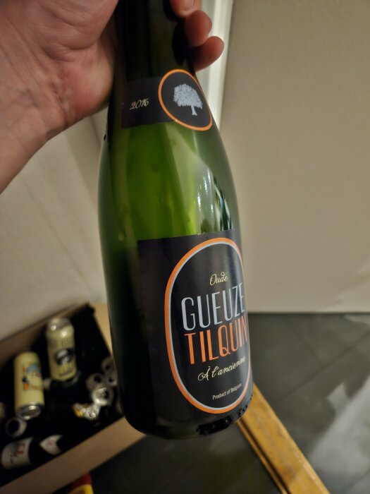 En hand håller en flaska Öude Gueuze Tilquin à l'ancienne. En ölflaska med etikett framför öllådor.