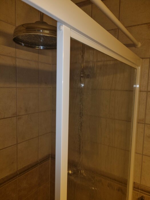 Hörn av duschutrymme, kakelvägg, duschhuvud, halvglansig glasdörr, badrumsinredning, beige kakel, metallstänger.