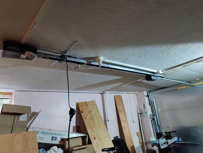Garageinteriör med öppen dörrmekanism, bråte, lådor, träplankor och hängande belysning under taket.