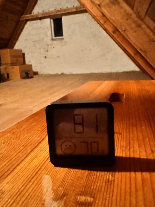 En inomhustermometer på ett trägolv i ett vindsvåningsrum med synliga takbjälkar och en liten fönster.