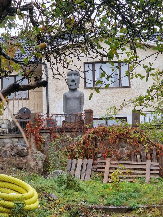 En stor stenstaty föreställande ett mänskligt ansikte i en trädgård framför ett hus.