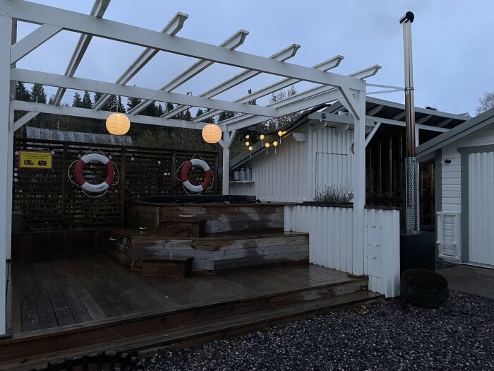 Utomhus bubbelbad med träbeklädnad, vita staket, ljusslingor, livbojar och en klarvitt pergola. Skymning eller gryning.