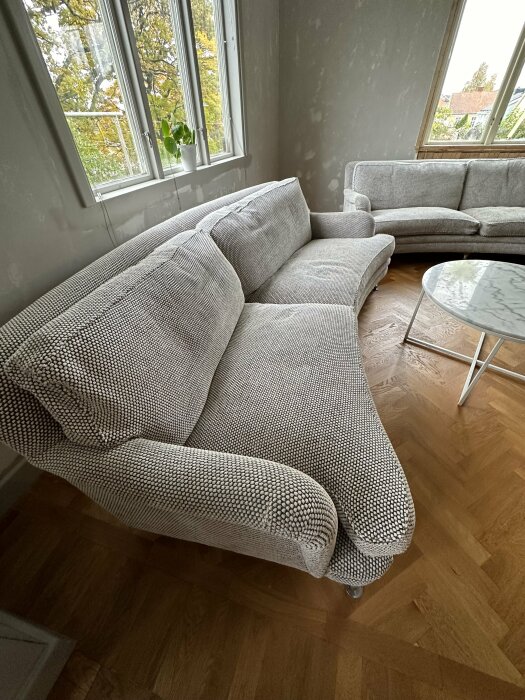 Vardagsrum med grå soffa, fåtölj, marmorbord, trägolv, fönster med grönska utanför.