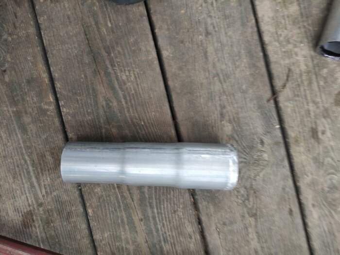 Silverfärgad cylinderformad föremål på trägolv. Otydligt, möjligen en metall- eller plastbit.