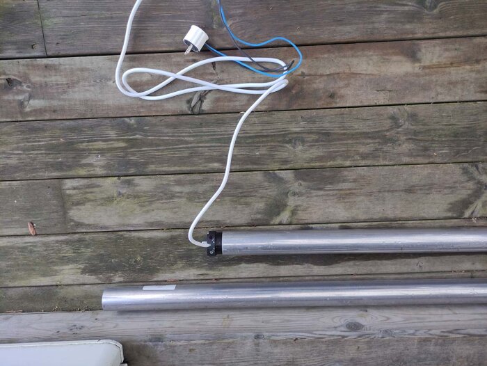 En vit mobiltelefonladdare på träaltan nära metallrör.
