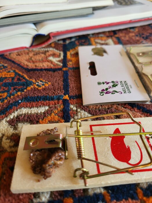 Musefälla med bete, öppen bok, och smartphone-förpackning på orientalisk matta.