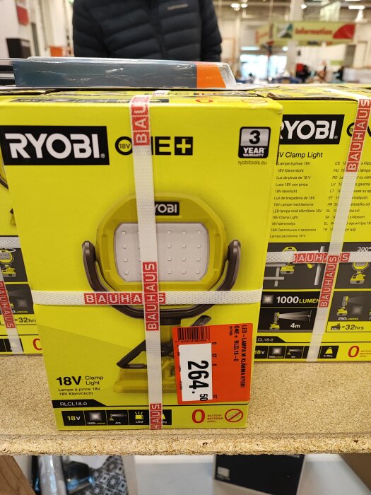 Ryobi 18V klämlampa i förpackning, BAUHAUS, pris etikett, 3-års garanti, bakgrund av butiksmiljö.