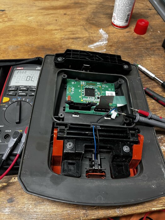 Elektronikenhet öppnad för reparation på träyta, multimeter, sprayburk och skruvmejsel synliga.