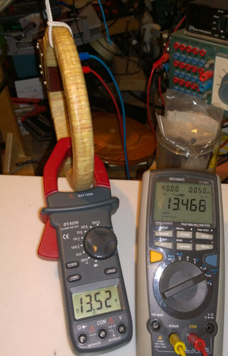 Experimentell uppställning med induktionsspole, strömtång, multimeter och elektronisk utrustning i bakgrunden.