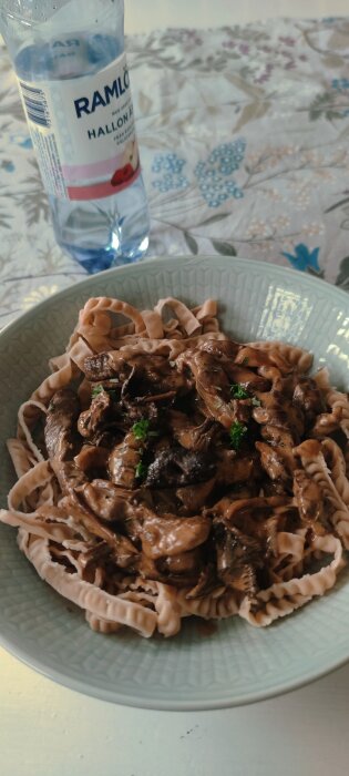 Pasta med svampsås på tallrik, flaska vatten med hallonsmak, blommigt dukunderlag, fönsterreflektion på bordet.