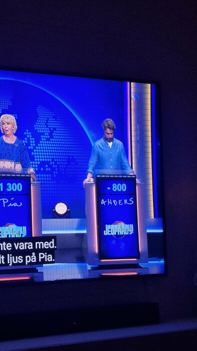 Skrämdump av Jeopardy-spel på TV. Två deltagare syns med sina poäng; en man står vid podium.