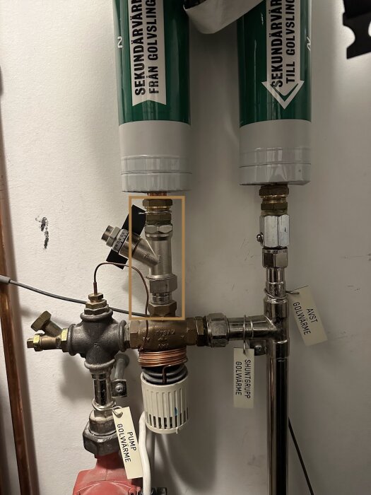 VVS-installation med rör, ventiler och etiketter märkta "Sekundärvatten från golvvärme" på en vägg.