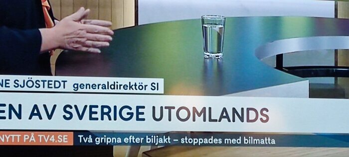 Person gestikulerar vid bord, glas vatten, bildtexter, nyhetsprogram, TV4-logotyp, grönt och blått bord.