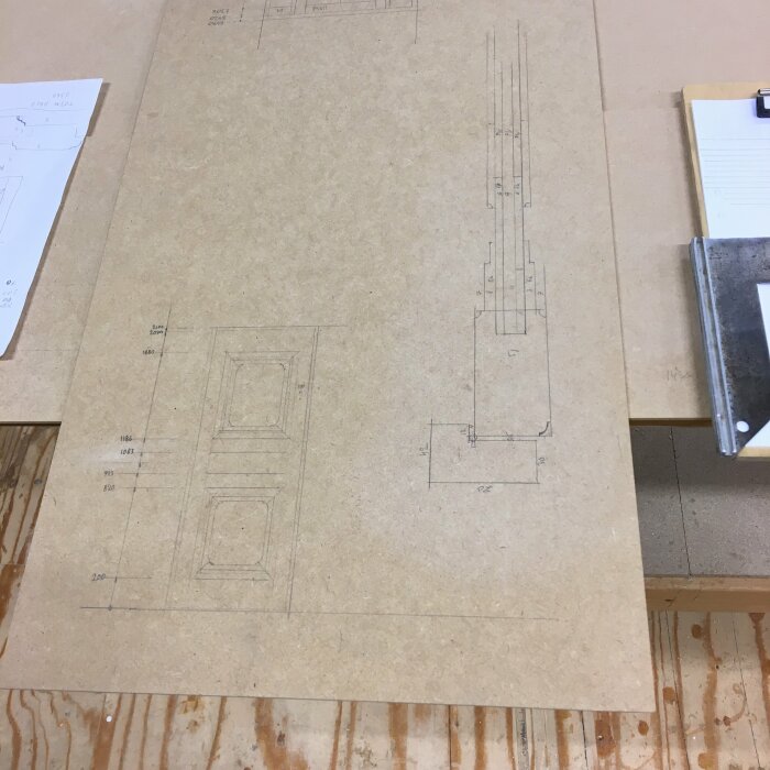 Arbetsyta med ritningar och mått för snickeri eller konstruktion, anteckningsblock och arbetsmaterial i verkstad.