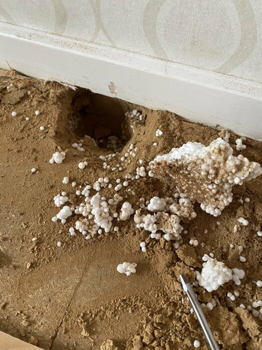 Hål i marken vid vägg, vita kulor, skum-lik substans, möjligt skadedjur eller isoleringsmaterial, skruvmejsel för skala.