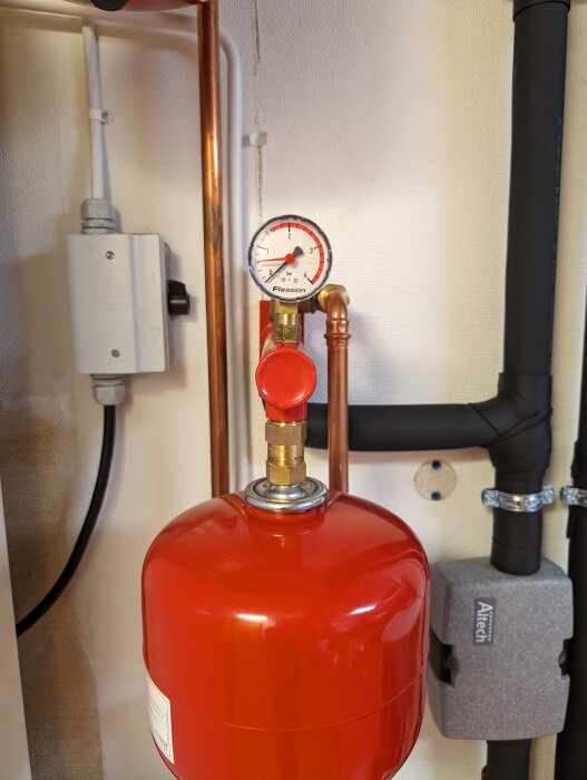Röd expansionskärl med manometer för vattentryck, kopparledningar, svart rör och elektriska komponenter, installation för uppvärmningssystem.