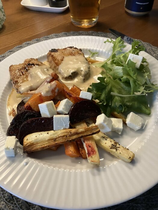 Middagstallrik med kycklingbröst, sås, rostade grönsaker, sallad och fetaost, serverat med öl.