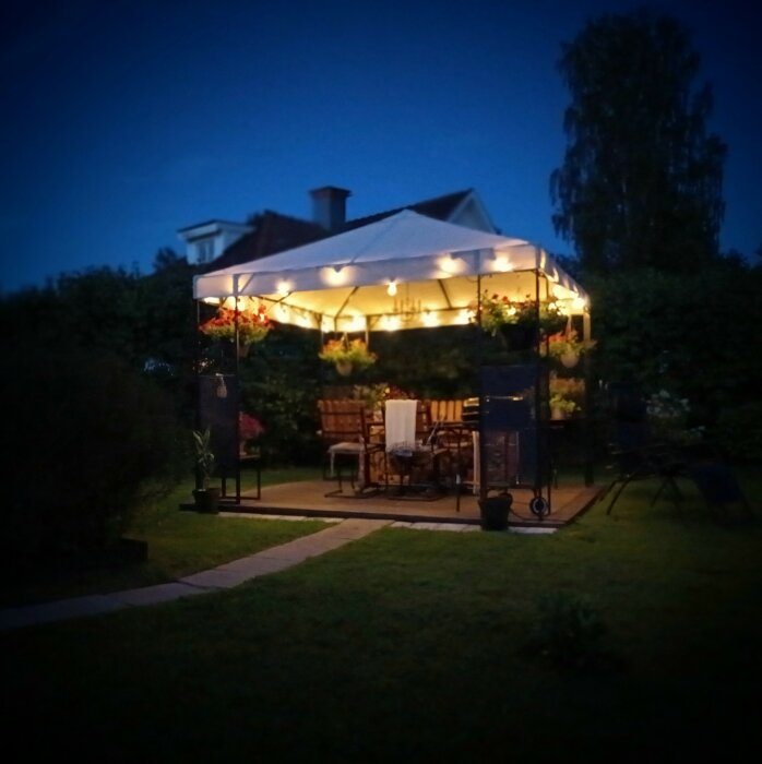 Kvällsträdgård med belyst paviljong, möbler och blomkrukor, mysig och inbjudande atmosfär.